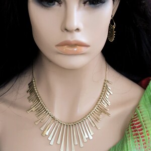 May include: A gold necklace with a geometric design. The necklace is made of small, thin metal pieces that are arranged in a fan shape. The necklace is worn by a mannequin with long black hair.