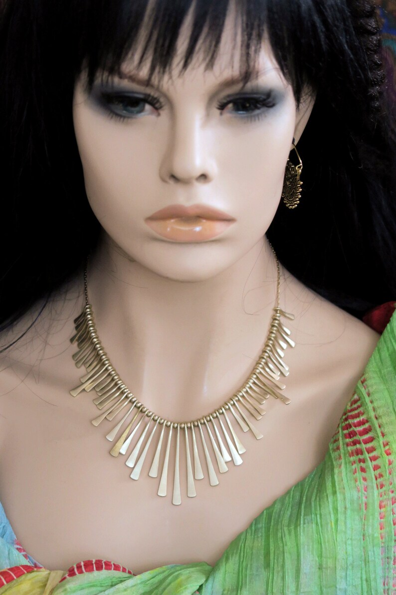 May include: A gold necklace with a geometric design. The necklace is made of small, thin metal pieces that are arranged in a fan shape. The necklace is worn by a mannequin with long black hair.
