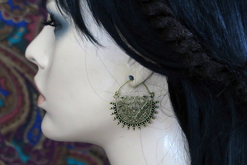 May include: A pair of gold-toned hoop earrings with a detailed, intricate design. The earrings are in the shape of a crescent moon with a floral pattern.