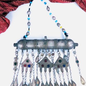 May include: A silver-toned metal amulet necklace featuring a cylindrical central tube with three triangular pendants hanging below. Each pendant is decorated with small red, blue, and green inset stones. The necklace has a beaded cord with silver, blue, white, and yellow beads.