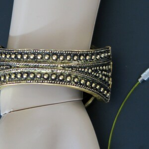 May include: A gold-toned metal bracelet with a beaded design. The bracelet is on a mannequin's wrist. A green cord with a silver clasp is in the background.