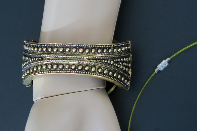 May include: A gold-toned metal bracelet with a beaded design. The bracelet is on a mannequin's wrist. A green cord with a silver clasp is in the background.