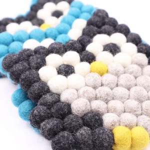 May include: A set of two colorful felt coasters with a geometric pattern. The coasters are made of small, round felt balls in shades of gray, white, yellow, and blue.