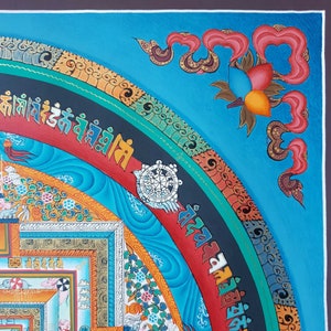 May include: A colourful Tibetan mandala painting with intricate details and a blue background. The mandala features a central wheel with spokes and a border of swirling patterns. The painting is framed with a dark brown border.