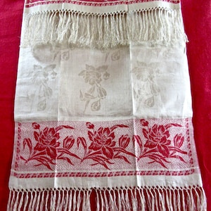 May include: A white linen tea towel with red floral embroidery and fringe. The towel has a damask pattern and is decorated with a repeating floral design.