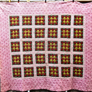 May include: A handmade quilt with a pink border and a central pattern of red and yellow squares. The squares are framed by green borders and arranged in a grid. The quilt's edges feature a wavy, patterned design.