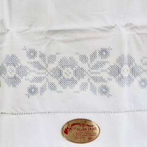 May include: A white cotton pillowcase featuring a blue cross-stitch floral pattern across the center. Below the embroidery, a horizontal hemstitch detail runs across the fabric. An oval gold label is attached, reading PILLOW CASES.