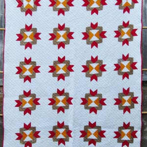 May include: A white quilt with a repeating pattern of brown, orange, and red starburst designs. The quilt has a red border and is hanging on a wooden structure. The quilt is a handmade item.