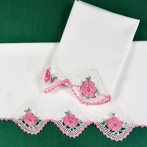 May include: Two white pillowcases with crocheted lace trim. The trim features pink roses and green leaves, with a scalloped pink border. One pillowcase is folded, and the other is unfolded, displaying the decorative edge.