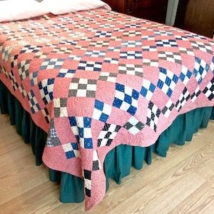 May include: A pink and white patchwork quilt with blue and black squares. The quilt is on a bed with a green bed skirt.