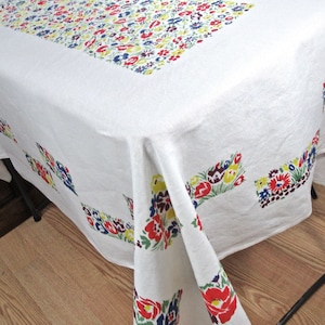 May include: White tablecloth with a floral pattern in red, yellow, blue, and green. The tablecloth has a border of smaller flowers and a larger floral design in the corners.