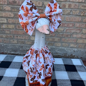 May include: A goose wearing a large bow and a skirt, both adorned with a repeating pattern of brown and orange bows on a white background. The goose's neck is white, and its beak is pink. The skirt has an orange and yellow trim.
