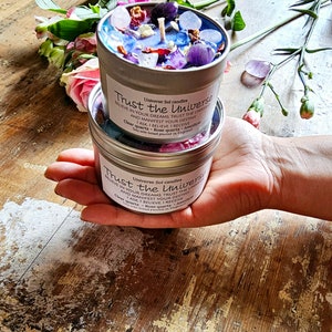 May include: A silver candle tin with a clear glass top filled with colorful crystals and a white candle. The tin has the text "Universe Sol candles Trust the Universe Believe in your dreams Trust the Universe and manifest your destiny I ask I believe I receive Clear quartz - Rose quartz - Amethyst Soy wax based poured in England" printed on it.