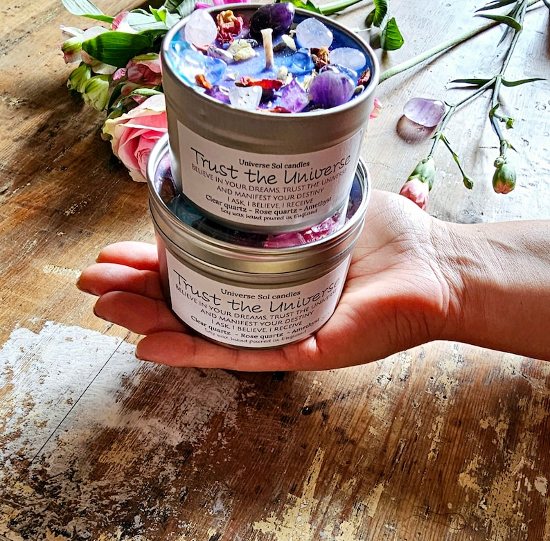 May include: A silver candle tin with a clear glass top filled with colorful crystals and a white candle. The tin has the text "Universe Sol candles Trust the Universe Believe in your dreams Trust the Universe and manifest your destiny I ask I believe I receive Clear quartz - Rose quartz - Amethyst Soy wax based poured in England" printed on it.