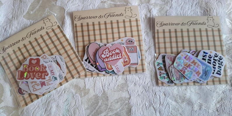 Może przedstawiać: Trzy zestawy naklejek o tematyce książkowej. Każdy zestaw ma inny wz&oacute;r, w tym naklejki z napisami "Book Lover", "Book Addict" i "In My Bookish Era". Naklejki znajdują się na beżowo-brązowym tle w kratkę.