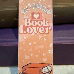 Może przedstawiać: Prostokątna zakładka do książki w kolorze brzoskwiniowym z napisem "SELF CONFESSED Book Lover". Ilustracja trzech ułożonych w stos książek. Tło w kolorze brzoskwiniowym z małymi sercami.