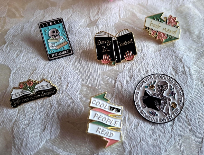 Może przedstawiać: Kolekcja emaliowanych przypinek z motywami książkowymi. Przypinki przedstawiają obrazy książek, czaszek i frazy takie jak "book nerd" i "Cool People Read". Paleta kolor&oacute;w obejmuje czerń, biel, złoto i r&oacute;żne odcienie niebieskiego i r&oacute;żowego.