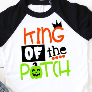 May include: White and black baseball style shirt with orange and green text that reads "King of the Patch" with a crown above the word "King" and a green pumpkin with a face below the word "Patch".