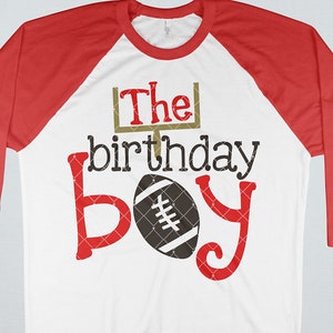 May include: A white and red baseball t-shirt with a football and the text "The birthday boy" in red and gold.