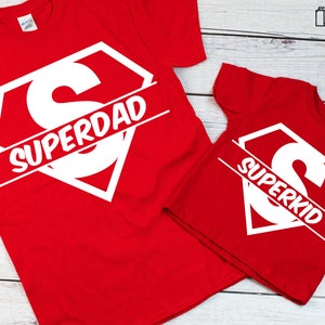 May include: Two red t-shirts with a white superhero logo and the words "Superdad" and "Superkid" printed on them.