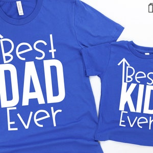 May include: Two blue t-shirts with white text. The larger shirt says "Best Dad Ever" with an arrow pointing up. The smaller shirt says "Best Kid Ever" with an arrow pointing up.