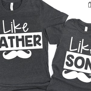 May include: Two grey t-shirts with white text and a white moustache graphic. The larger shirt says "Like Father" and the smaller shirt says "Like Son".