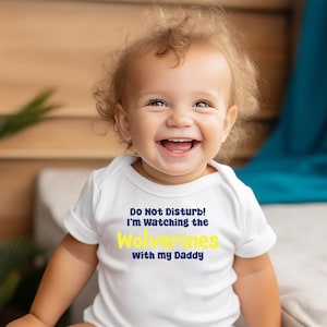 May include: A white baby onesie with a blue and yellow graphic that says "Do Not Disturb! I'm Watching the Wolverines with my Daddy."