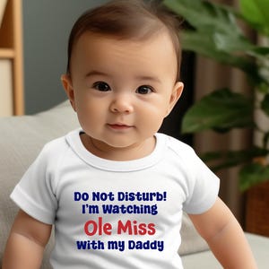 May include: A baby wearing a white onesie with the text "Do Not Disturb! I'm Watching Ole Miss With my Daddy" in blue and red lettering. The baby is sitting on a light gray couch in front of a green plant.