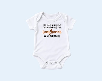 May include: A white short-sleeved baby bodysuit featuring the black text "Do Not Disturb! I'm watching the" followed by the word "Longhorns" in brown, and "With my Daddy" in black. The garment has a round neckline and three metal snaps at the base.
