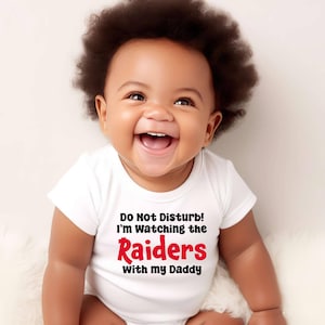 May include: A white baby onesie with the text "Do Not Disturb! I'm Watching the Raiders with my Daddy" in black and red lettering. A baby with dark curly hair is wearing the onesie and smiling.