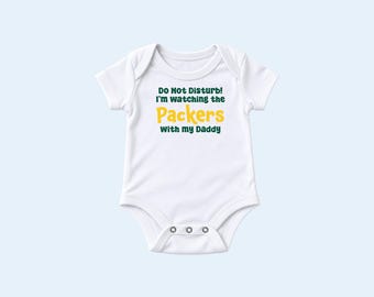 May include: A white short-sleeved baby bodysuit featuring the dark green text "Do Not Disturb! I'm watching the" followed by the yellow text "Packers" and the dark green text "with my Daddy" printed on the center of the chest area.