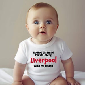 May include: A white baby onesie with the text "Do Not Disturb! I'm Watching Liverpool with my Daddy" in black and red lettering.