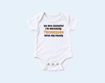 Peut inclure: Un body pour bébé blanc à manches courtes avec le texte noir "Do Not Disturb! I'm watching", le mot "Tennessee" imprimé en orange, suivi de "With my Daddy" en noir. Le vêtement présente une encolure ronde et trois boutons-pression métalliques.