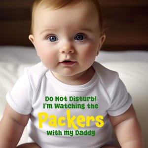 May include: A baby wearing a white onesie with the text "Do Not Disturb! I'm Watching the Packers with my Daddy" in green and yellow letters.