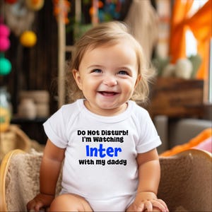 May include: A baby wearing a white onesie with the text "Do Not Disturb! I'm watching Inter with my daddy" in black and blue lettering. The baby is smiling and looking at the camera.