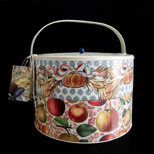 May include: A round, two-tiered tin with a white handle and lid. The top tier features a repeating pattern of blue diamonds and pumpkins with ribbons. The bottom tier displays apples and pears. A small tag hangs from the handle.