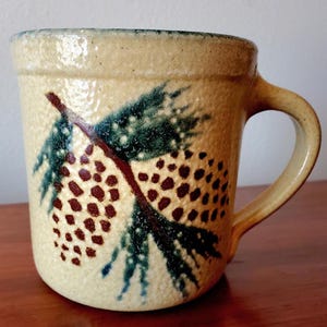 May include: A ceramic mug with a brown, green, and white pine cone design. The mug has a textured surface and a handle.