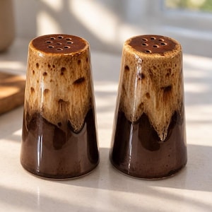 May include: A pair of ceramic salt and pepper shakers. The shakers are cone-shaped with a dark brown base that fades into a lighter brown and tan speckled glaze. Each shaker has a series of holes at the top for dispensing.