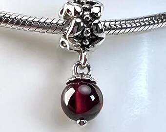 May include: A silver charm with a floral design and a dangling, round, deep red bead. The charm is attached to a silver bracelet. The bead has a polished, reflective surface.