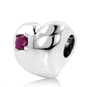 May include: A polished silver heart-shaped charm with a vibrant red gemstone. The charm has a smooth, reflective surface and a small, round hole. The gemstone is set in a square, textured setting.