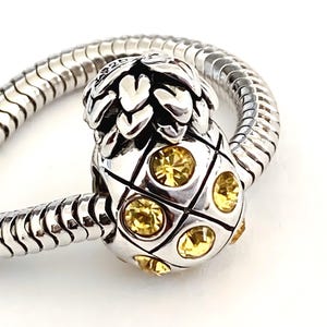 May include: A silver pineapple charm with yellow gemstones, attached to a silver snake chain bracelet. The charm has a detailed crown and a grid pattern. The bracelet is a classic design, suitable for various styles.