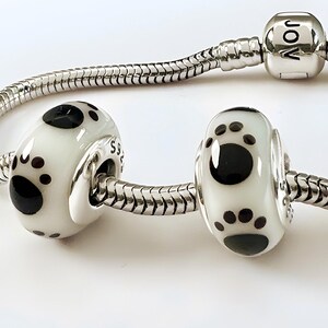 K&ouml;nnte beinhalten: Zwei wei&szlig;e und schwarze Pfotenabdruck-Charm-Perlen an einem silbernen Schlangenkettenarmband. Die Perlen haben eine gl&auml;nzende Oberfl&auml;che mit einem gro&szlig;en schwarzen Pfotenabdruck und kleineren schwarzen Punkten. Das Armband hat einen silbernen Verschluss.