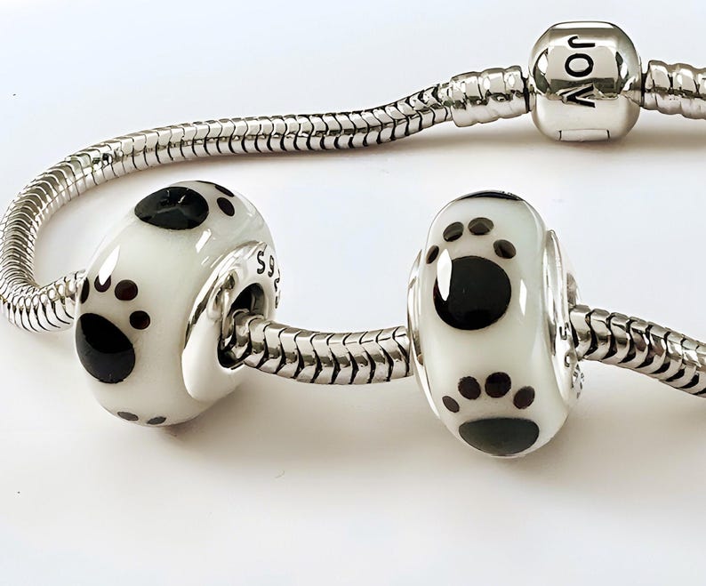 K&ouml;nnte beinhalten: Zwei wei&szlig;e und schwarze Pfotenabdruck-Charm-Perlen an einem silbernen Schlangenkettenarmband. Die Perlen haben eine gl&auml;nzende Oberfl&auml;che mit einem gro&szlig;en schwarzen Pfotenabdruck und kleineren schwarzen Punkten. Das Armband hat einen silbernen Verschluss.