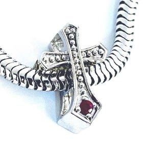 May include: A silver-toned metal charm shaped like a cross with a beaded texture along its edges. The cross features a single, small, round, deep red gemstone set near the bottom point. The charm is threaded onto a flexible, segmented silver chain.