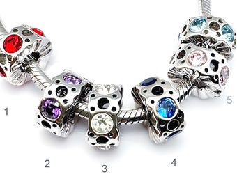 May include: A selection of silver charm beads with coloured gemstones. The beads are round with a patterned design and feature red, purple, clear, blue, and pink stones. The beads are strung on a silver chain.