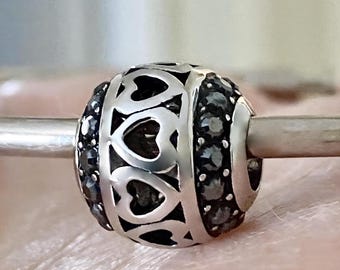 May include: A spherical charm bead featuring a central band of cut-out heart shapes in polished silver-tone metal, flanked by two rows of small, dark, faceted stones. The bead is threaded onto a thin, smooth, silver-tone metal rod or bracelet wire.