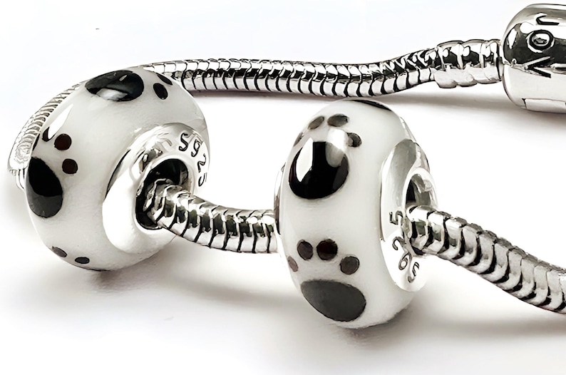 May include: Close-up of a silver bracelet with two white and black paw print beads. The beads have a glossy finish and are accented with silver details. The bracelet is a stylish accessory.