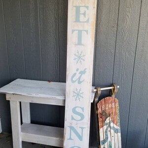 Puede incluir: Un letrero de madera blanco con las palabras "Let it Snow" pintadas en azul claro. El letrero est&aacute; decorado con copos de nieve y est&aacute; apoyado en una mesa de madera blanca. Un trineo vintage con un mu&ntilde;eco de nieve pintado en &eacute;l est&aacute; al lado del letrero.