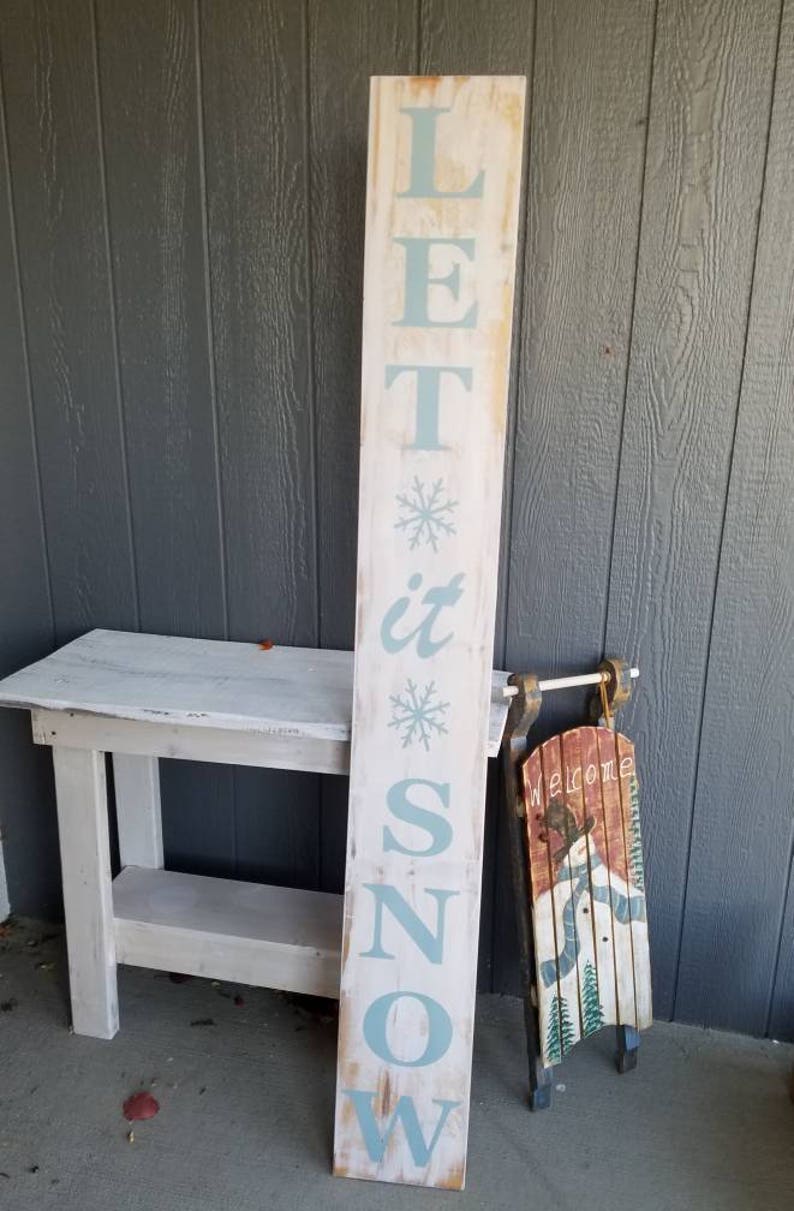Puede incluir: Un letrero de madera blanco con las palabras "Let it Snow" pintadas en azul claro. El letrero est&aacute; decorado con copos de nieve y est&aacute; apoyado en una mesa de madera blanca. Un trineo vintage con un mu&ntilde;eco de nieve pintado en &eacute;l est&aacute; al lado del letrero.