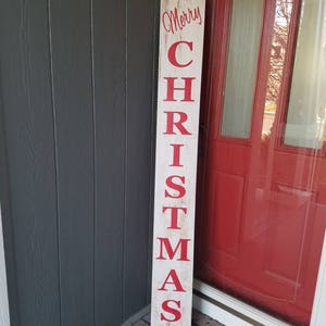 Puede incluir: Un letrero de madera blanco con las palabras "Merry Christmas" en letras rojas. El letrero est&aacute; apoyado contra una puerta roja.